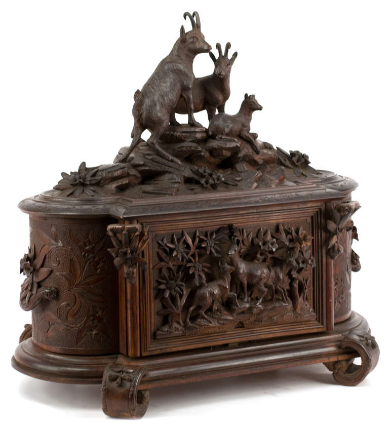 The exterior is ornately carved linden wood with full-relief sculptures of mountain goats on the front and top. The lid lifts to reveal a mirrored interior with removable drink service with four cut-glass liquor bottle and six cut-glass and gilt