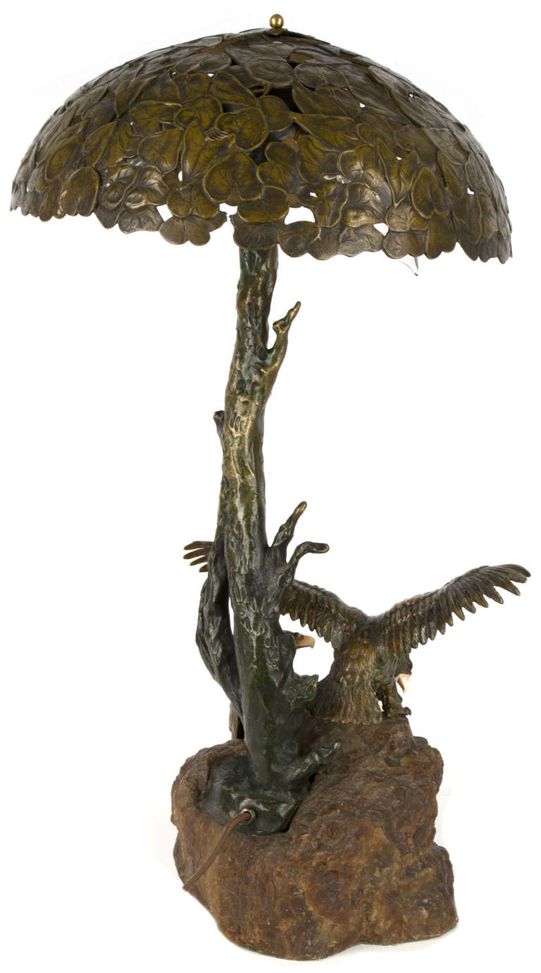 An exquisitely sculpted Austrian bronze table lamp featuring two large eagles with bone beaks standing on a rocky outcrop —Â made of onyx — under the shade, itself a bronze sculpted canopy of leaves upheld by a bronze tree trunk.
