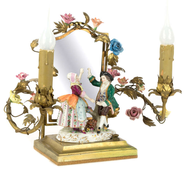 A pair of elaborate boudoir table lamps with porcelain figures and flowers, inset into ormolu vinery and placed against a bevelled  glass mirror.