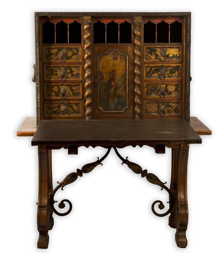 A Spanish drop-front desk, trimmed and decorated in iron beadwork, leather, and red velvet. The door opens downward, providing a writing surface, to reveal painted scenes of animal life, brass lion-head pulls, and Portuguese-turned columns (i.e.