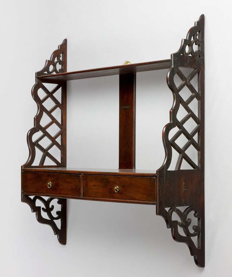 A fine set of George III Chippendale period mahogany hanging shelves; having pierced fretwork sides and with two drawers which retain their original axehead handles. 

THOMAS CHIPPENDALE (1718-1779)
