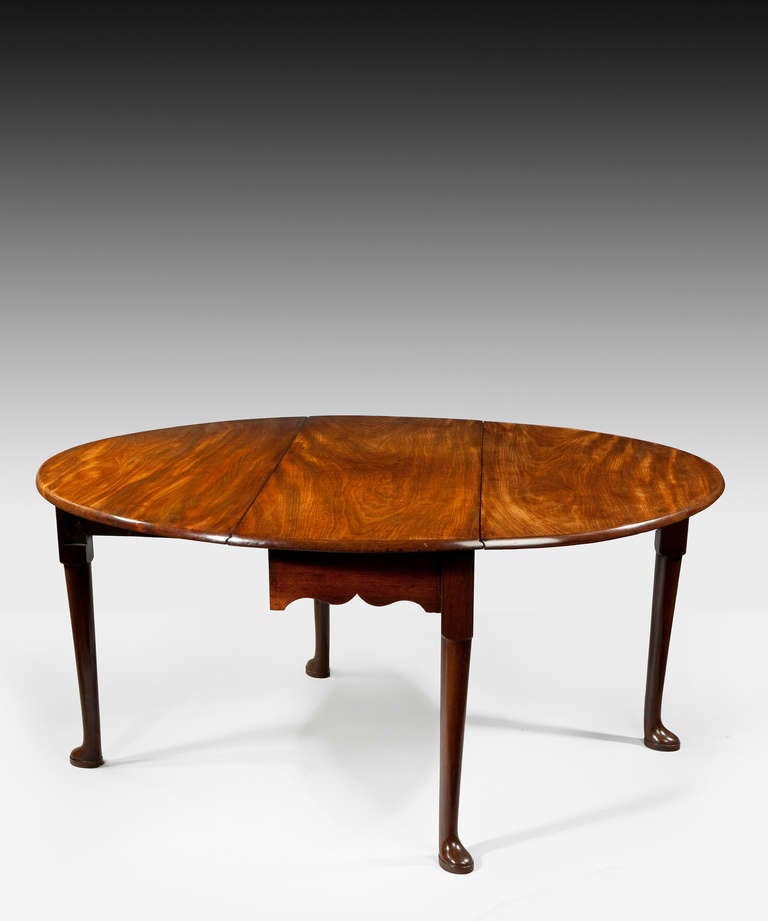 A George II period mahogany oval drop-leaf dining table; the well figured top above an ogee arch shaped frieze; raised on club legs which terminate in pad feet. This dining table retains a beautiful slightly faded color. This dining table's