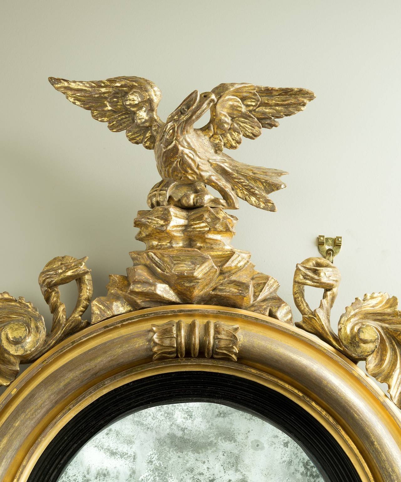 A Regency period carved giltwood convex mirror; the original convex mirror plate set within a reeded ebonised slip and a carved giltwood frame which is decorated with lotus leaves; the frame is surmounted by an eagle flanked by snakes standing on a