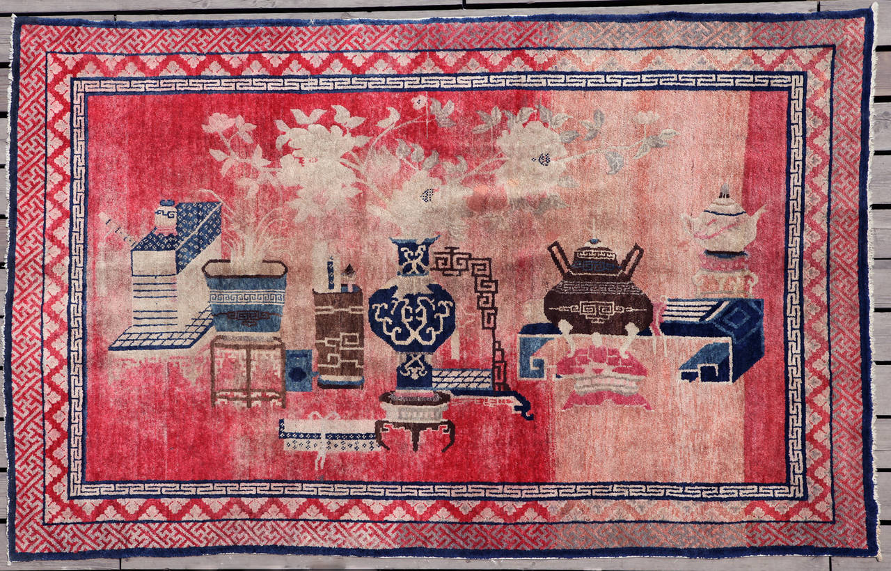 This stunning Inner Mongolian pictorial rug combines warm red hues with an intriguing pattern. 

Made for the kang, the Northern Chinese platform bed, this rug depicts the symbols of the Four Accomplishments (chessboard, lute, paintings and books)