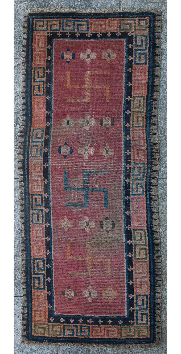 This very boldly drawn monastic rug has three large swastikas, meaning auspicious in Sanskrit, decorating the central field. 
The combination of the swastikas with the red background, the colour traditionally associated with the Tibetan clergy,