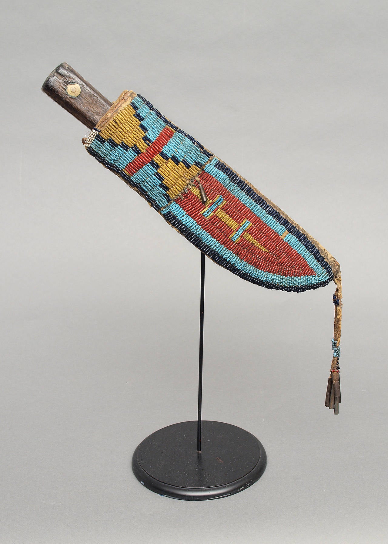 This sheath is constructed of native tanned hide with red, yellow, light and dark blue trade beads and sinew sewn. Geometric designs down the front panel, finished with a partially beaded bifurcated suspension with tin cone danglers. Created by a