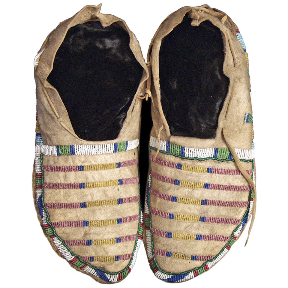 A vintage/antique pair of Native American beaded moccasins, created circa 1870 during the Late Classic Period of North American Indian art by a Crow (Plains Indian) artist. Composed of native tanned hide and partially beaded in a classic banded