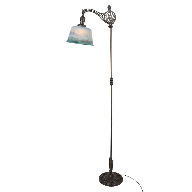 The ornate cast bridge bracket and pedestal bring plenty of drama to this early 20th century floor lamp, but the hand-painted glass shade brings all the charm. The scene depicts a tiny seaside village, and has been painted on the inside of the