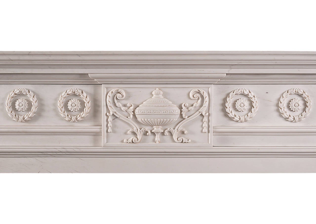 A late Georgian style carved fireplace in white marble, the centre tablet with an ornate urn with stylised scrolled decoration either side, and Vitruvian scrolls around the urn, the top scrolled jambs with oval floral decoration. Moulded shelf. A