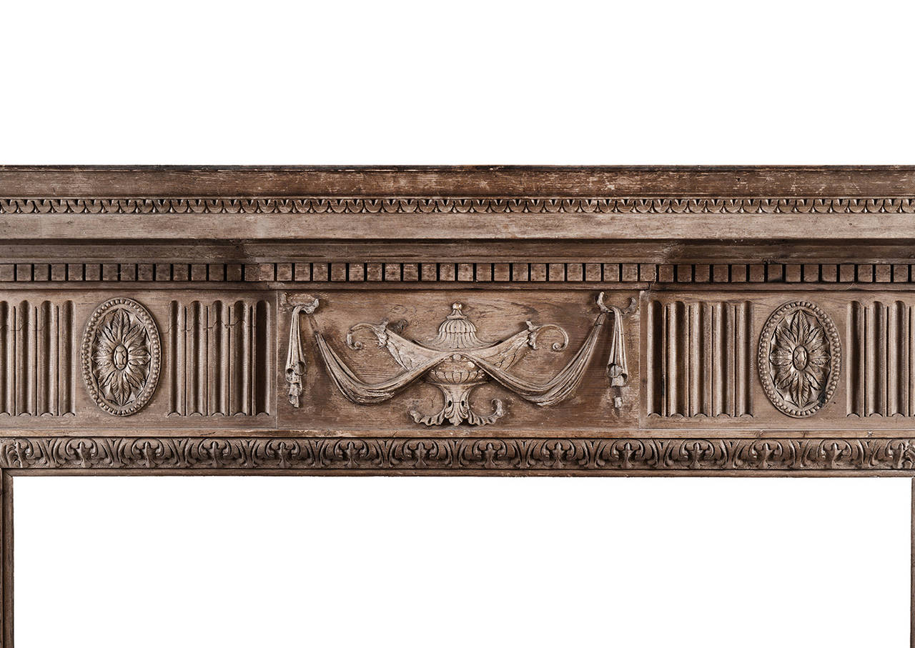 An English neoclassical pine fireplace. The centre blocking with carved urn and drapery, flanked by oval paterae and fluting. The tapering legs with tied ribbons and bellflowers, with Greek key detailing to base. Carved acanthus leaves to inner