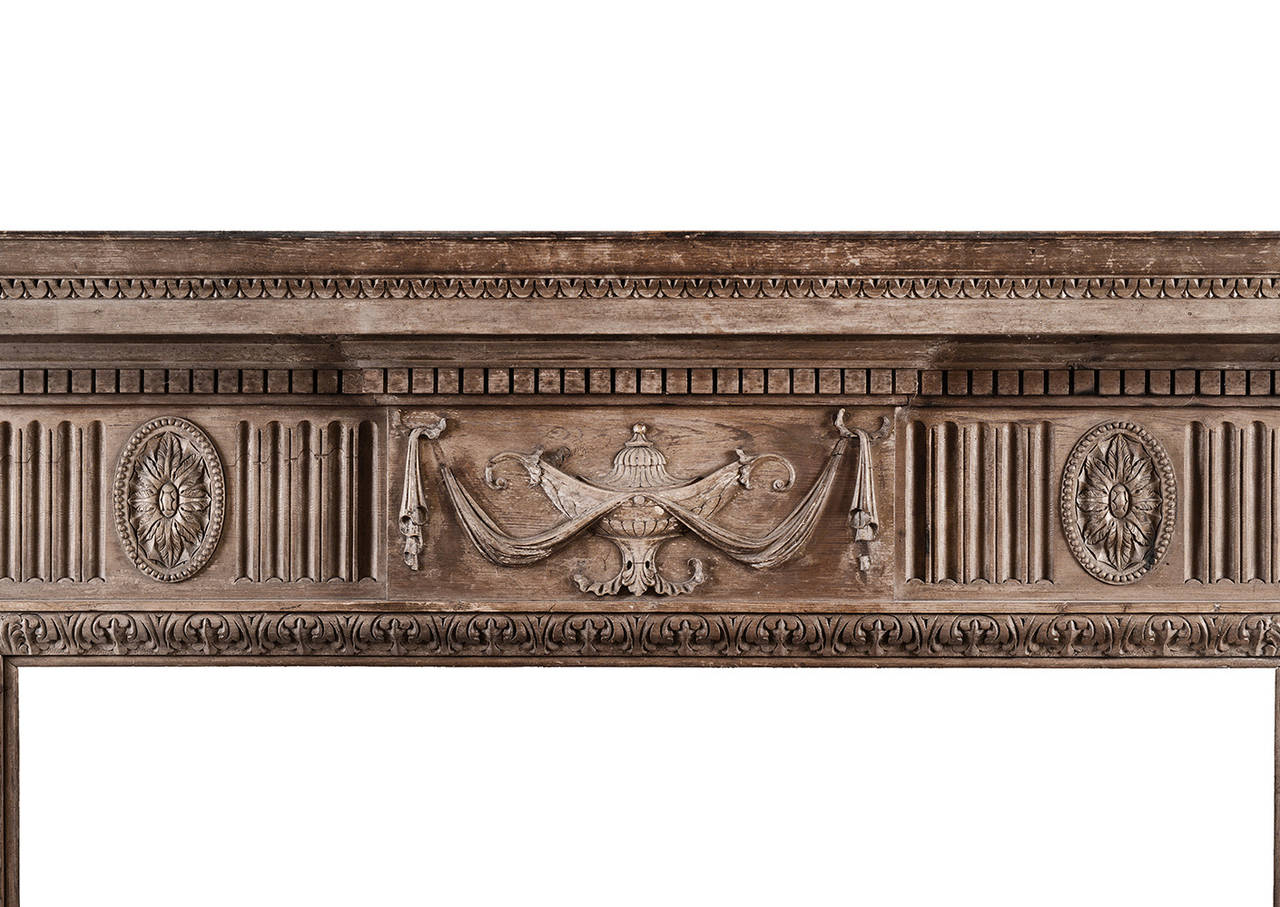 Une cheminée anglaise néoclassique en pin. Le bloc central avec urne et draperie sculptées, flanqué de paterae ovales et de cannelures. Les pieds effilés sont ornés de rubans noués et de campanules, et la base est ornée d'une clé grecque. Feuilles