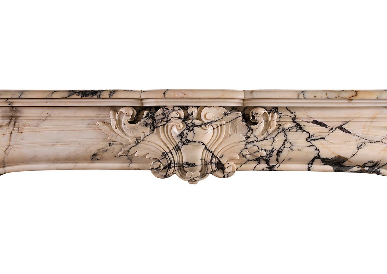 A French antique fireplace in the Louis XIV / Louis XV transitional style, made from Italian Pavonazzo marble. The arched frieze with carved cartouche and foliage to centre flanked by shaped panels and scrollwork. The shaped, panelled jambs with