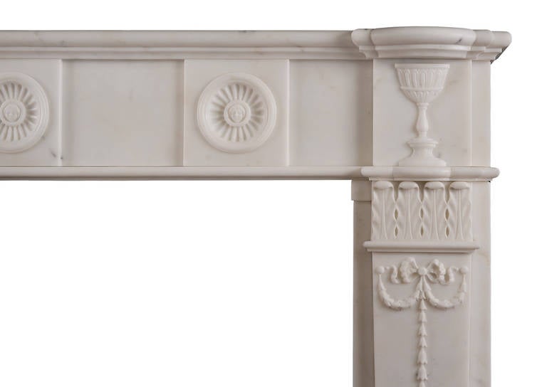 A carved English antique fireplace in statuary marble. The jambs with tapering pilasters featuring finely carved bell drops and drapery surmounted by stiff acanthus leaf capitals. The frieze with concaved fluted paterae with carved mask to centre