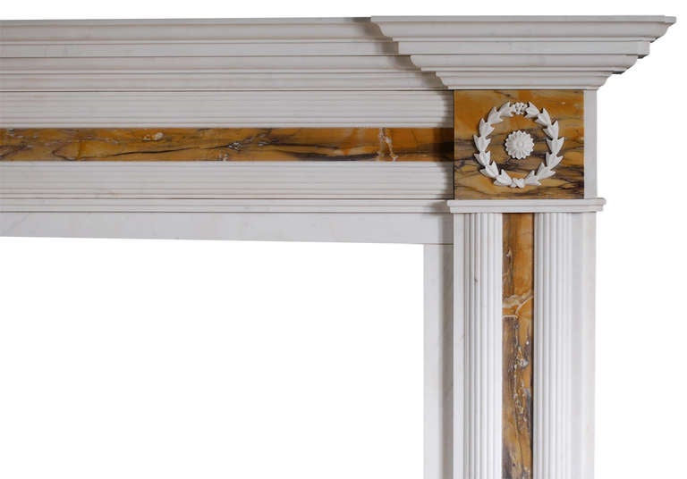 Une cheminée anglaise de style Régence en marbre blanc avec incrustation de Sienne. La frise et les jambages sont ornés de roseaux demi-ronds, les blocs d'extrémité de rosettes délicates entourées de guirlandes sculptées. Étagère moulée en façade.