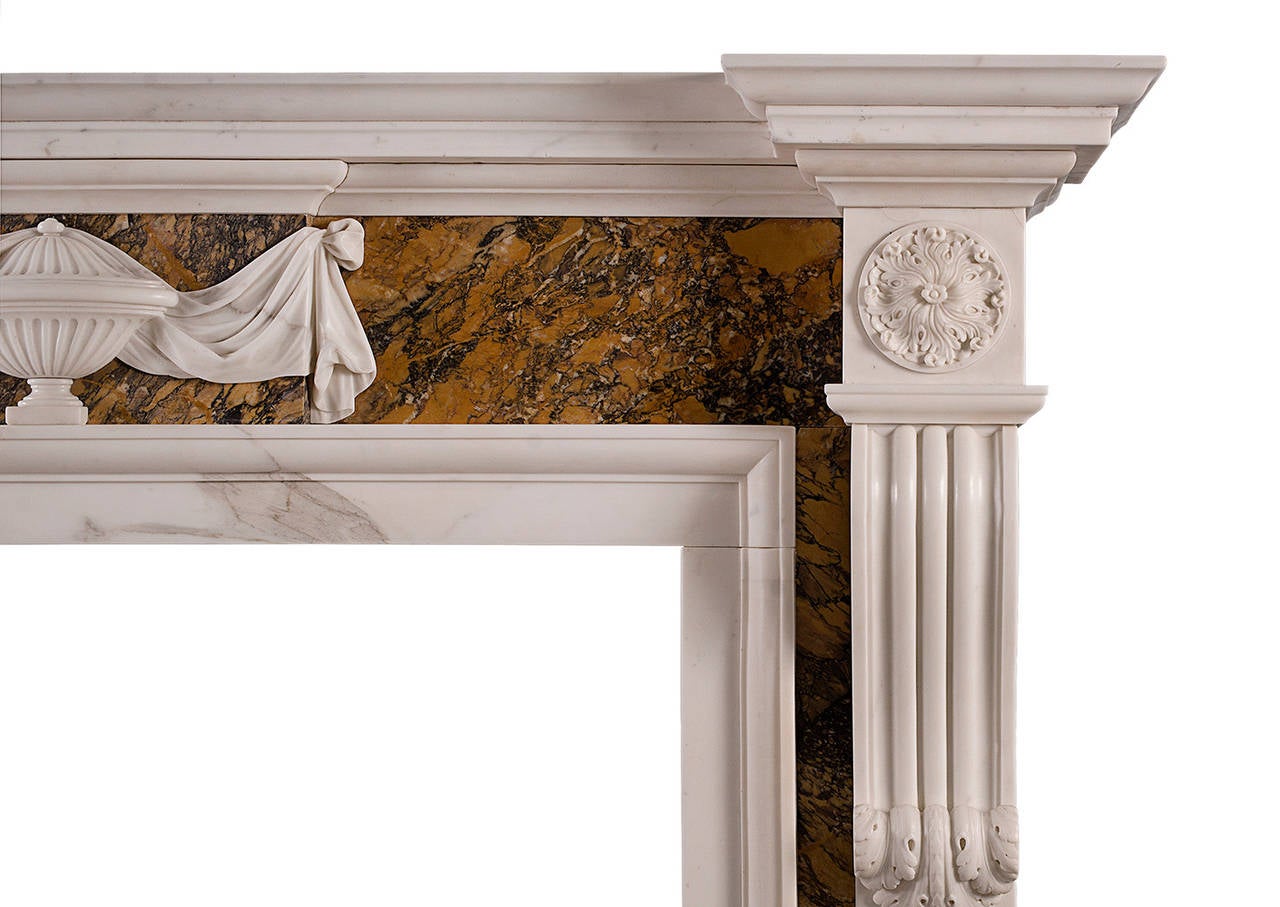 An English, Georgian style fireplace in statuary marble with Siena inlay. The panelled jambs with carved brackets, surmounted by carved end blockings. The Siena marble frieze with centre blocking featuring urn with cascading drapery. Moulded shelf