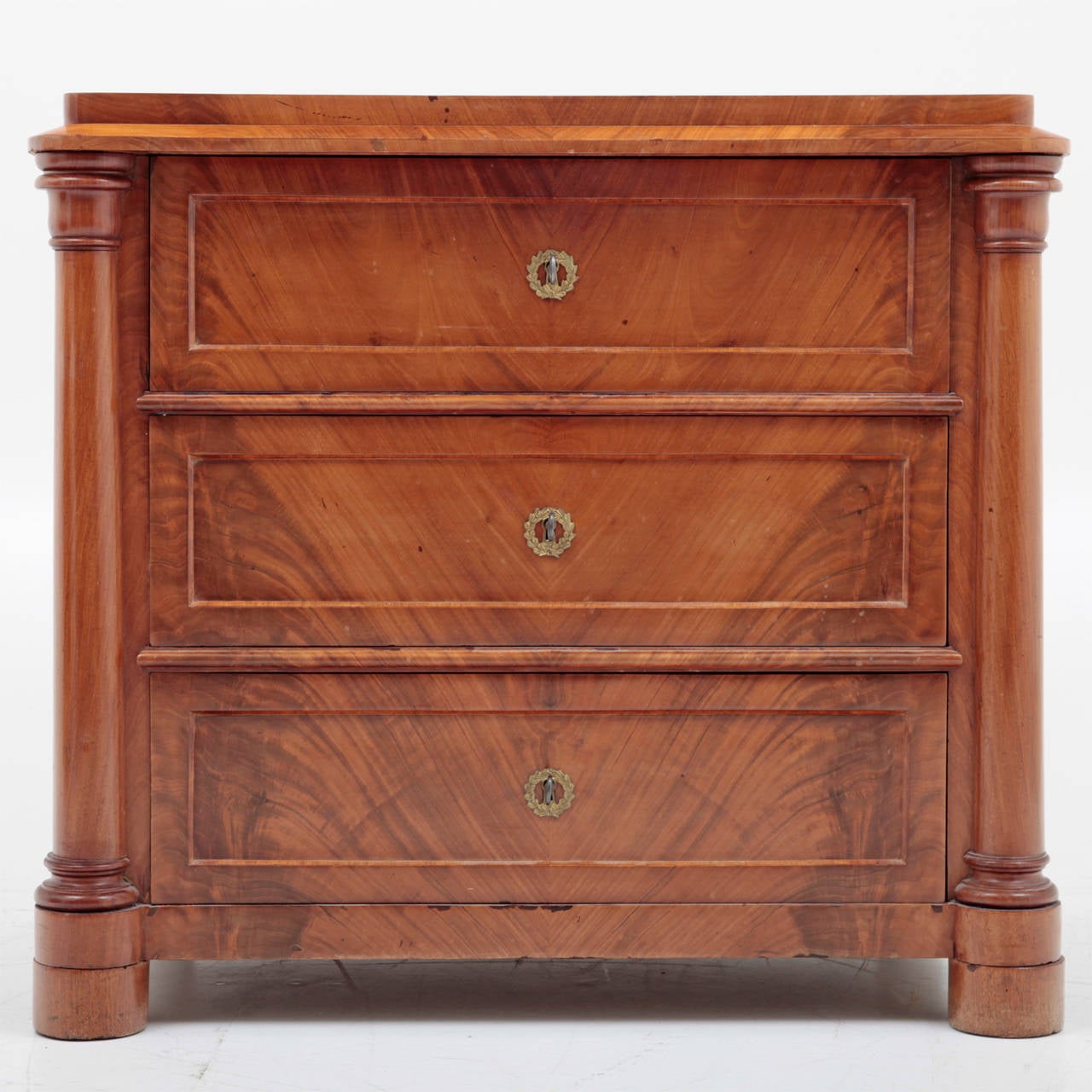 The chest has three drawers, each with a coffered front and round brass escutcheons. The front two feet are cylindrical, the rear two are square. The front corners are decorated with three-quarter columns. The upper edge is slightly protruding while