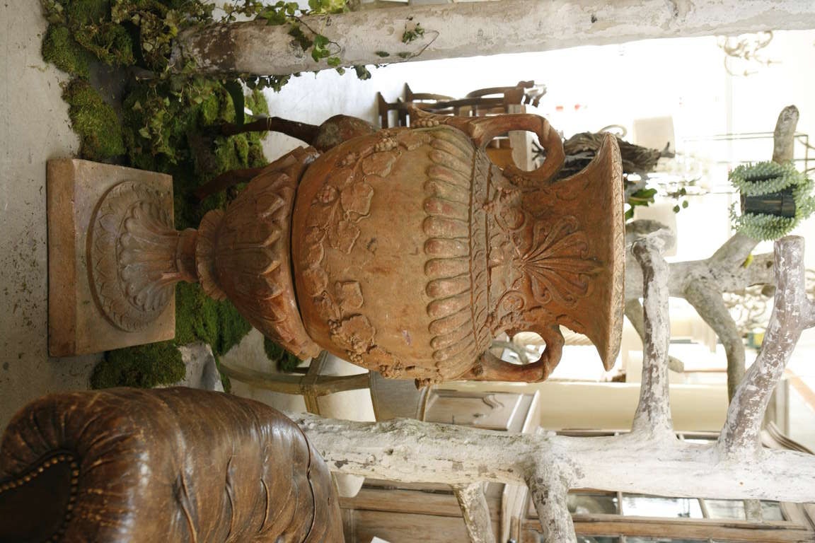 A single large red terra-cotta urn with floral decoration sitting on a pedestal. Exposure to the elements from being outdoors has caused beautiful patina.