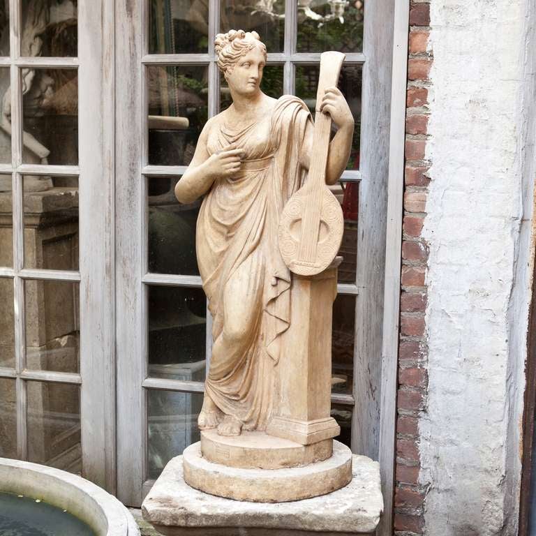 This beautiful terra cotta statue, representing Euterpe, (Greek Muse of music & lyric poetry) features the muse facing left, resting on a raised squared plinth and holding an upright musical instrument. The whole raised on a round cast stone base.