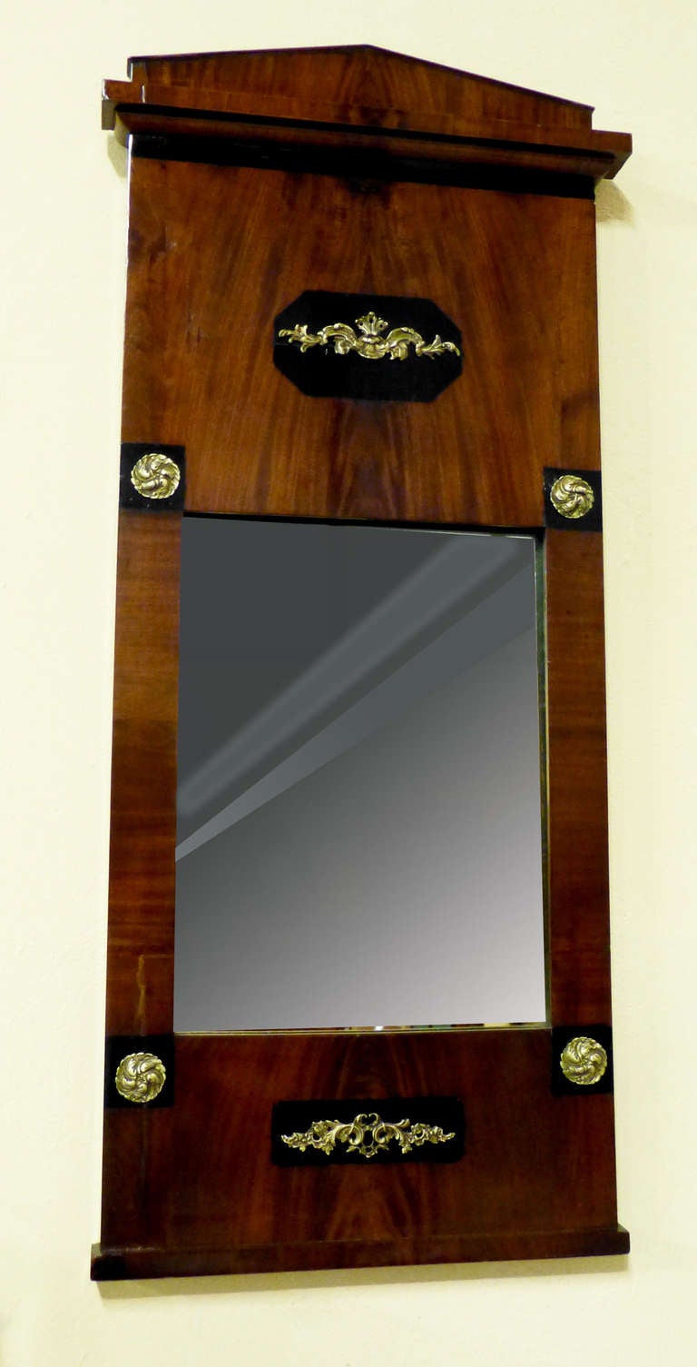 Figured mahogany late Empire early 19th century  Biedermeier mirror bearing Empire gilt brass decorations over ebonised backrounds. The pediment is of the Schinkel type similar to  Greek Doric, over flat mouldings and base. The mahogany is finely