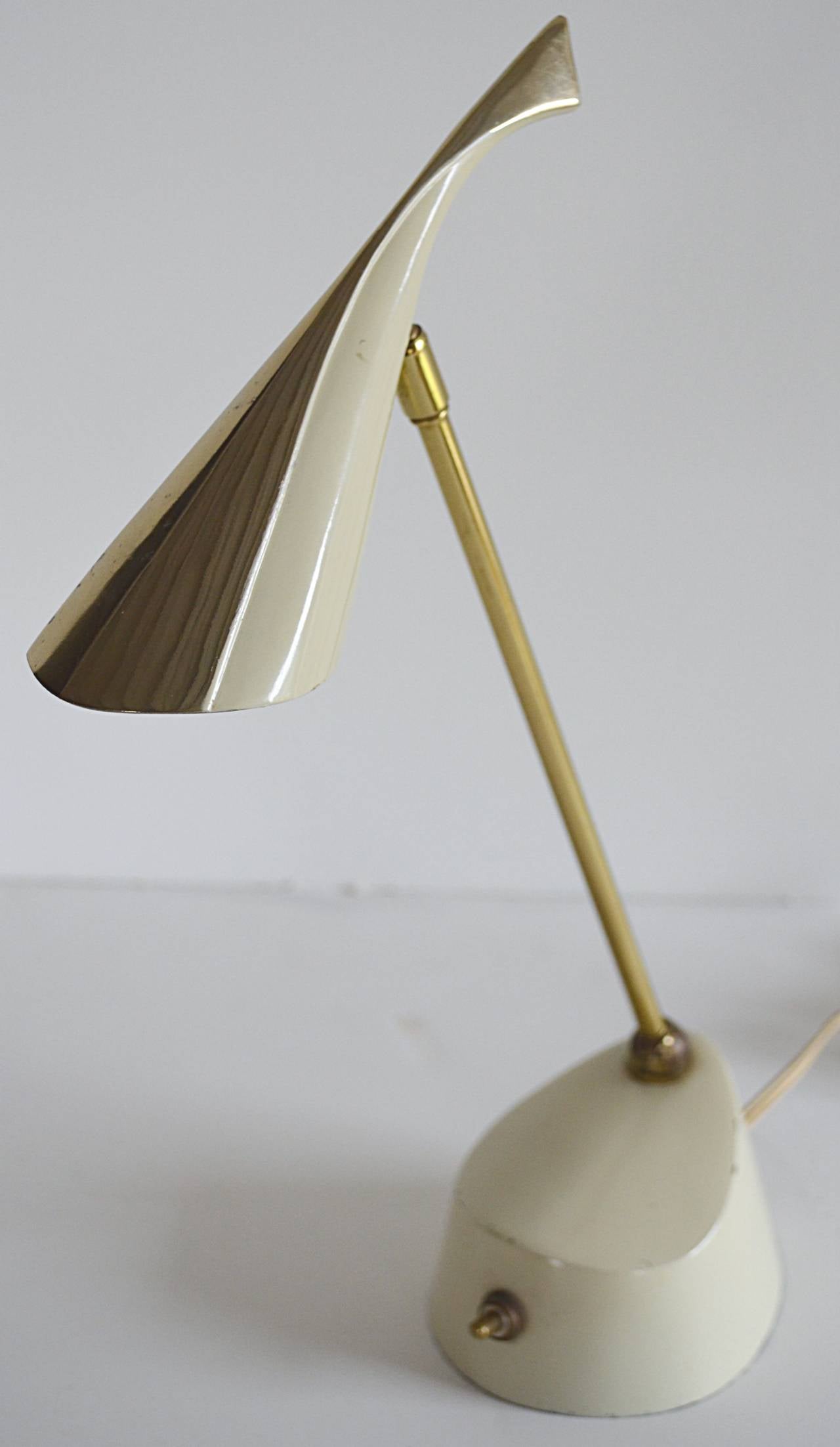 This little gem can be used as a desk lamp or hang on the wall as a sconce. The bone white enameled weighted base supports a brass pivoting shaft and pivoting biomorphic shade in brass and bone enamel. Original wiring.
