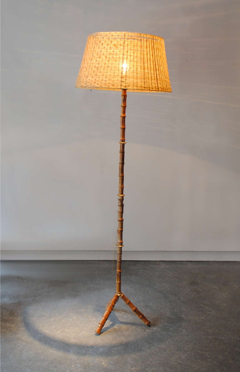 A mid-century modern floor lamp by Jacques Adnet in bamboo and bronze rings with its original rafia shade. There are slight imperfections to the shade. The lamp has been rewired.