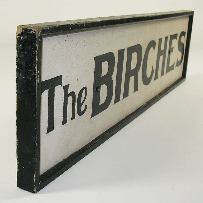 A framed solid wood sign painted white with black hand lettering that served as the name marker for a cottage among birch trees.