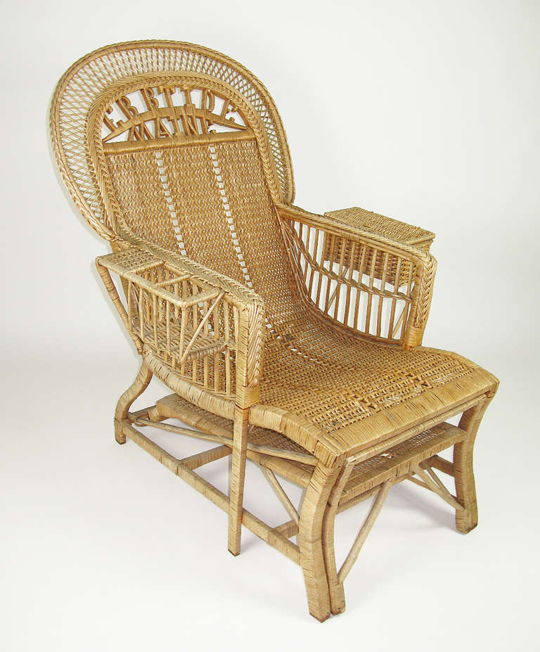 This wicker chair has a retractable footrest, along with a drink holder and storage basket on the arm rests. Wicker lettering within the back arch says 