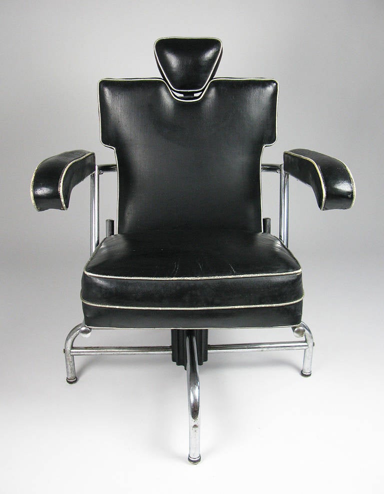 This modernist chair has a tubular chromed steel frame and black faux leather upholstery with silver contrast piping. The headrest adjusts to numerous heights, and the chair swivels and changes height upon full rotations. It is an extremely heavy