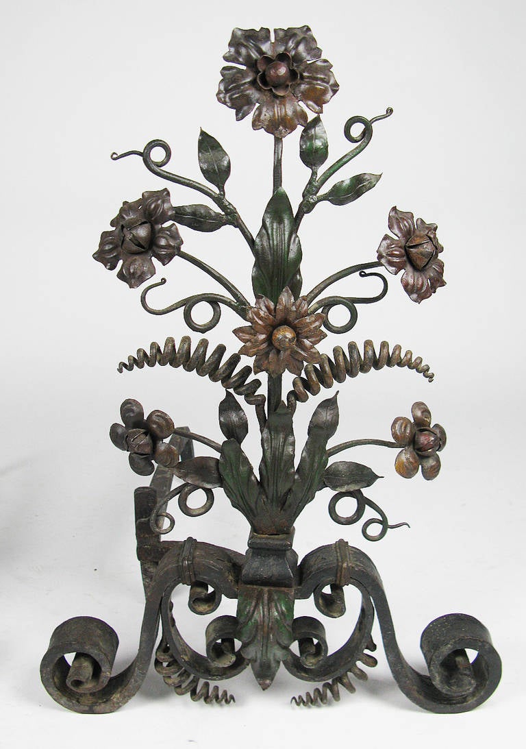 These figural, wrought-iron andirons have an elaborate floral, foliate and spiral tendril design, with a sturdy scrolled iron base. They retain traces of their original polychrome paint decoration in shades of green, yellow/orange and red, which