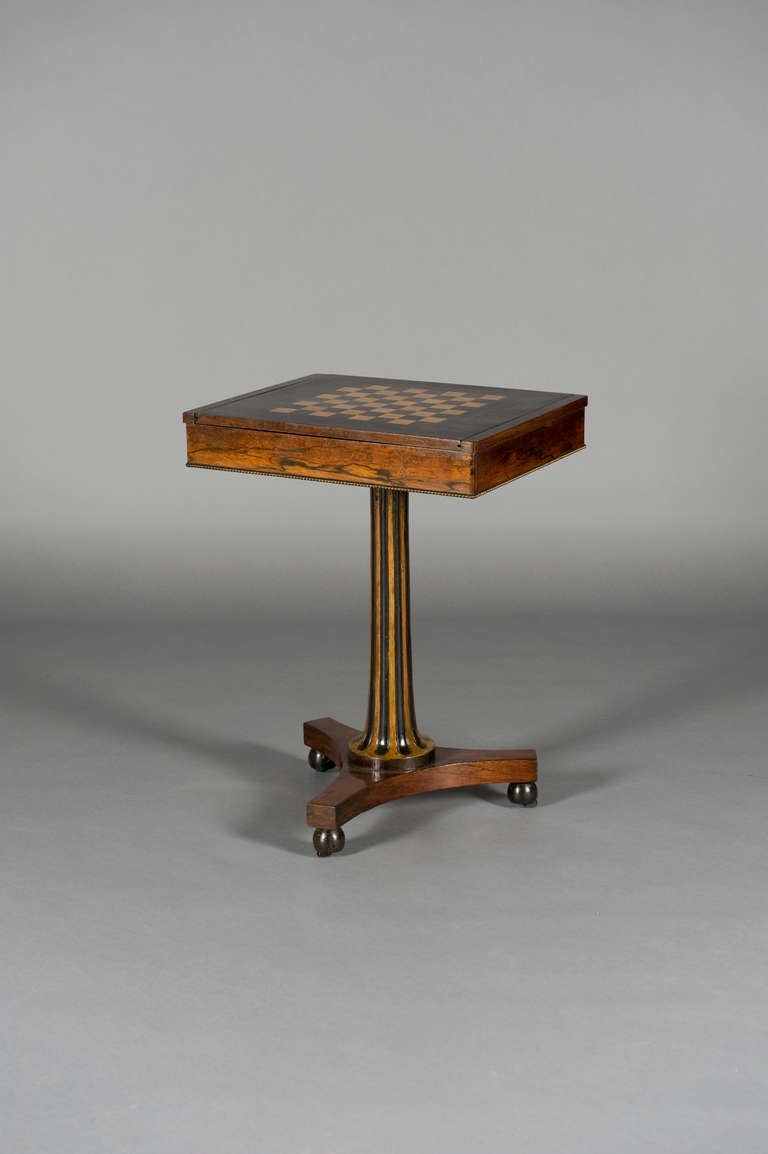 The boxed top having a reversable slide to reveal an inlaid chess board and well for chess pieces. Edged with beaded brass moulding. Standing on a simulated rosewood reeded and gilded column, on a triform base with turned feet conceiling castors. In