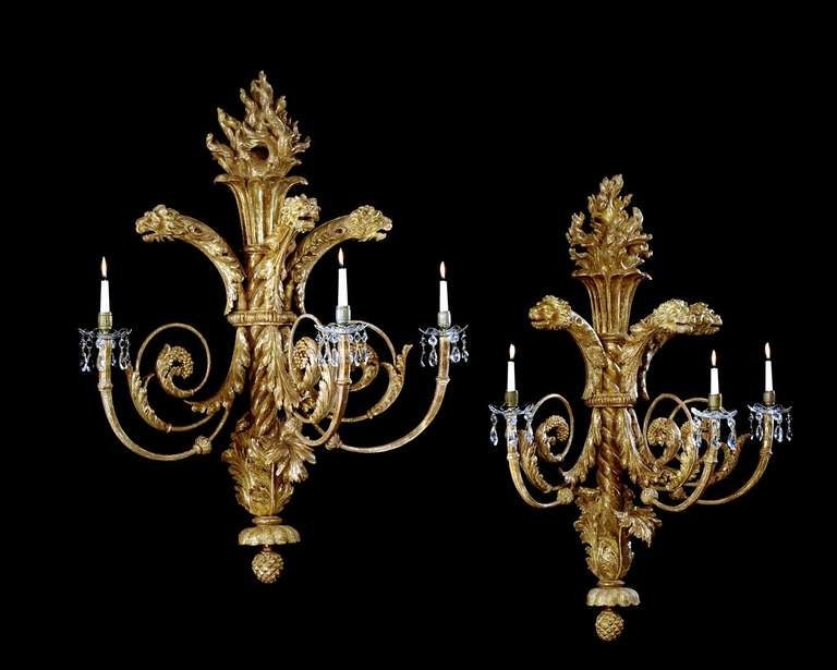 An impressive and large-scale pair of early 19th century carved giltwood wall lights in the form of flaming torches with triple-scrolled acanthus and berry carved arms with brass socles and cut-glass drip-pans hung with drops, surmounted with carved