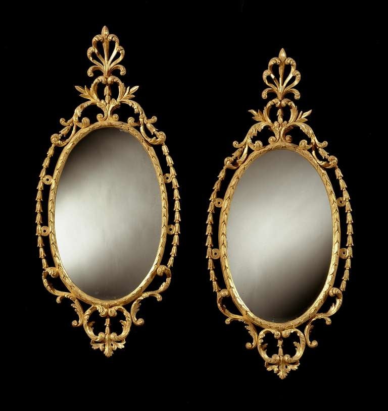A pair of late 18th century carved and giltwood oval mirrors with later 19th century plates surrounded by a laurel leaf border and an outer border of husks, with a scrolled apron and a foliate cresting surmounted by palmettes.