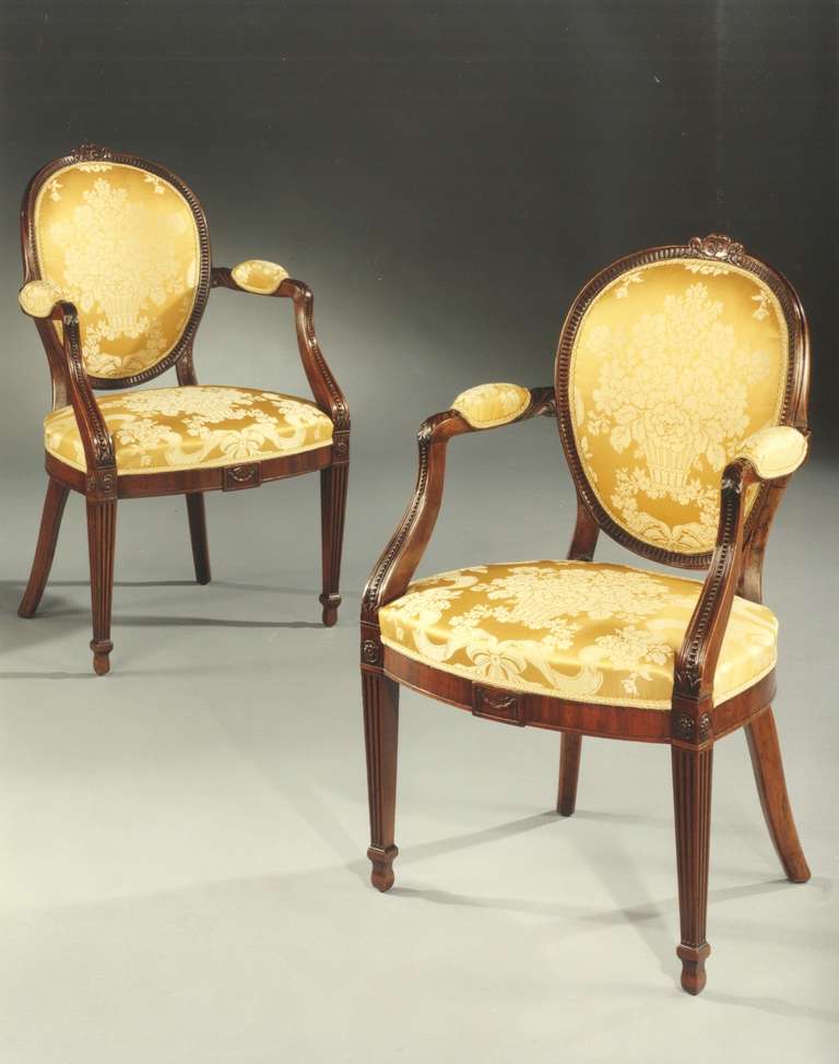 A pair of late 18th century Adam period mahogany armchairs attributed to Gillows of Lancaster, each having an oval stuffed back with fluted mahogany border and cresting with finely carved leaf and husk swags, and outswept padded arms with downswept