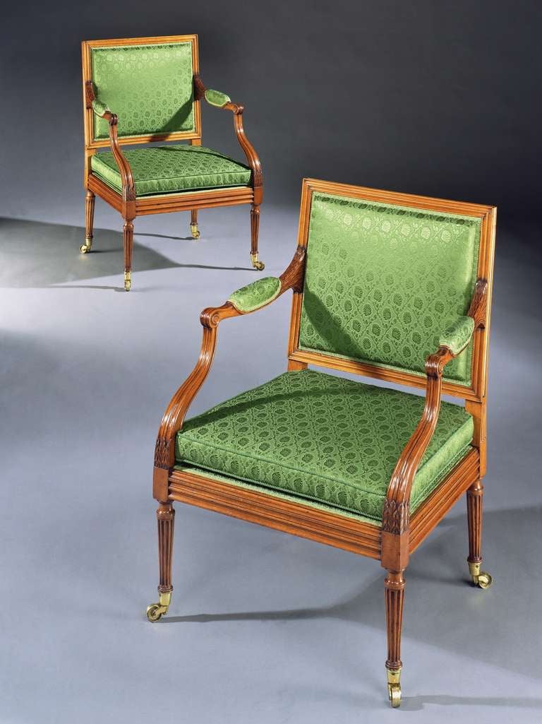 An unusual pair of late 18th century carved satinwood armchairs each with a channelled square framed rectangular padded back and squab cushion covered in green silk damask, with padded down swept reeded arms with scroll terminals, above a channelled