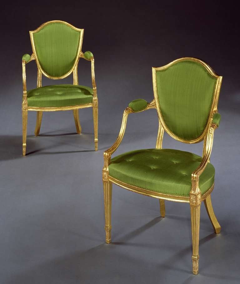 A fine pair of late 18th century carved giltwood Hepplewhite period shield back armchairs, the moulded frames with stuffed backs and seats covered in float buttoned green silk; on square fluted tapering legs headed by paterae.