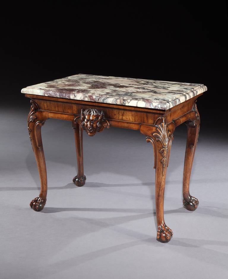 A mid 18th century carved walnut side table attributed to Giles Grendey, having an 18th century replaced brèche violette marble top above a cross veneered concave frieze applied with a central satyr mask; on cabriole legs with crisply carved