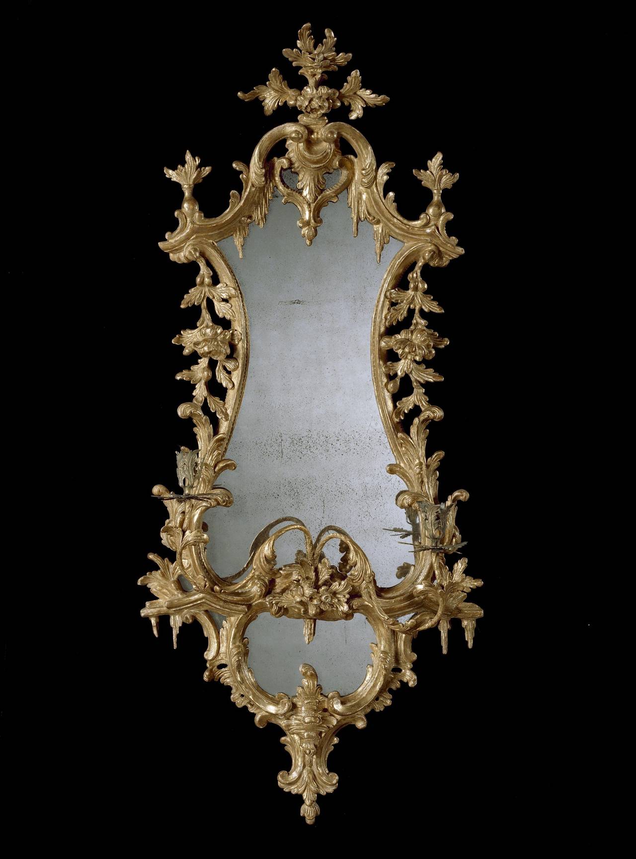 A pair of mid-18th century Chippendale period carved giltwood girandoles, each having 18th century replaced mirror plates within a waisted giltwood frame with shaped canopy carved with waterfall acanthus leaf decoration and trailing flowers to the