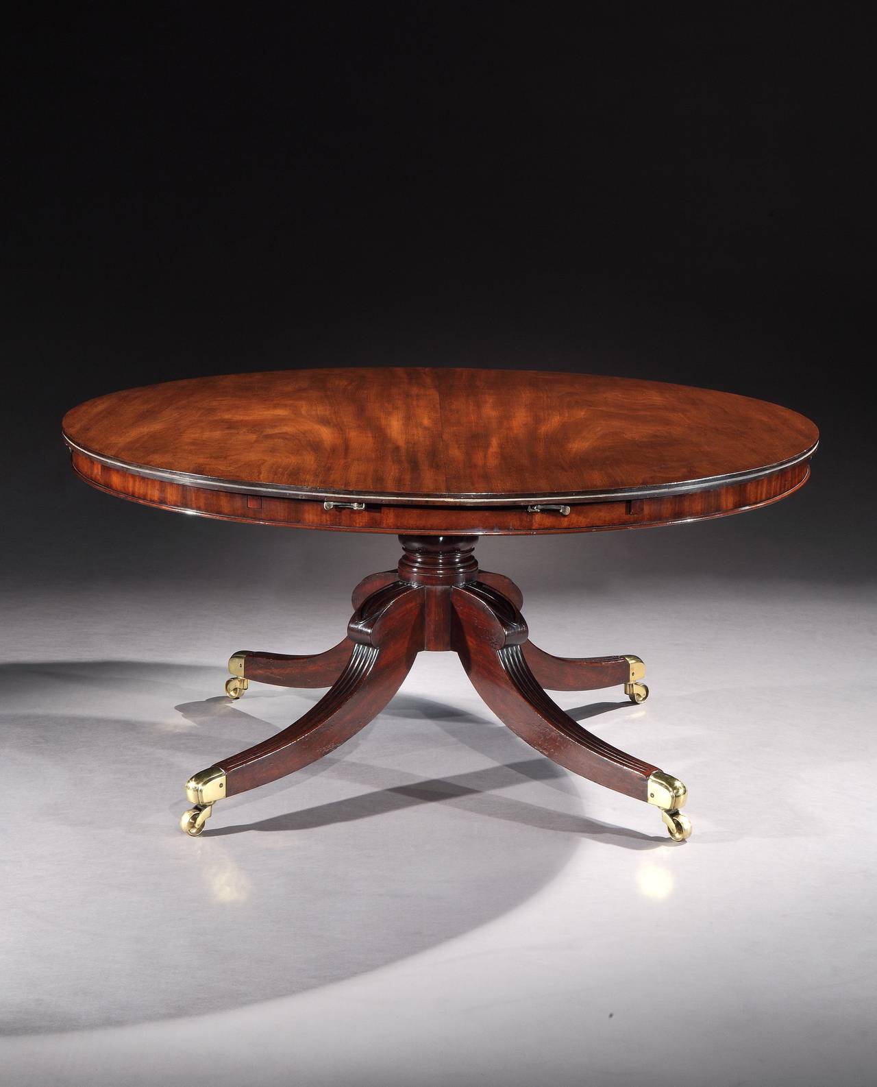 An early 19th century circular extending dining table retaining the original quarter round extending leaves, having a tip-up top supported by a turned baluster column with four reeded, hipped splay legs terminating in the original brass cap