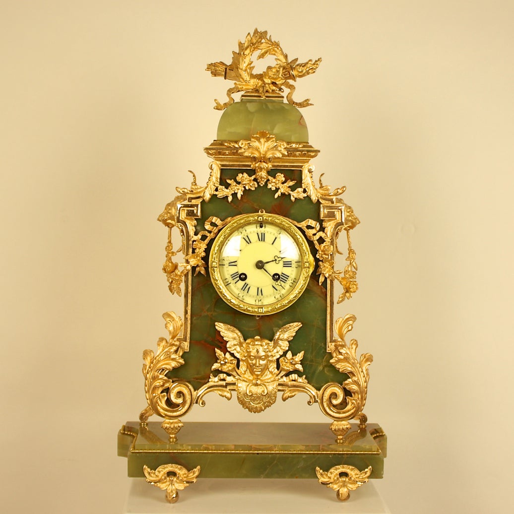 Onyx mantel clock of cartouche shape, containing a pale yellow porcelain dial with Roman and Arabic numerals. The case surmounted by a dome shaped roof mounted with ribbon tied wreath and torches. The surrounding onyx case framed with gilded
