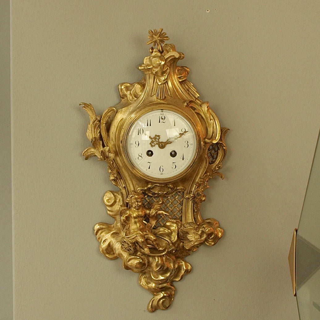 The white enamel dial with Arabic numerals, contained within a cartouche-shaped case surmounted by a star, rocks and C-scrolls, flanked by acanthus leaves, the base cast with a winged cupid in a chariot pulled by two pigeons riding on clouds