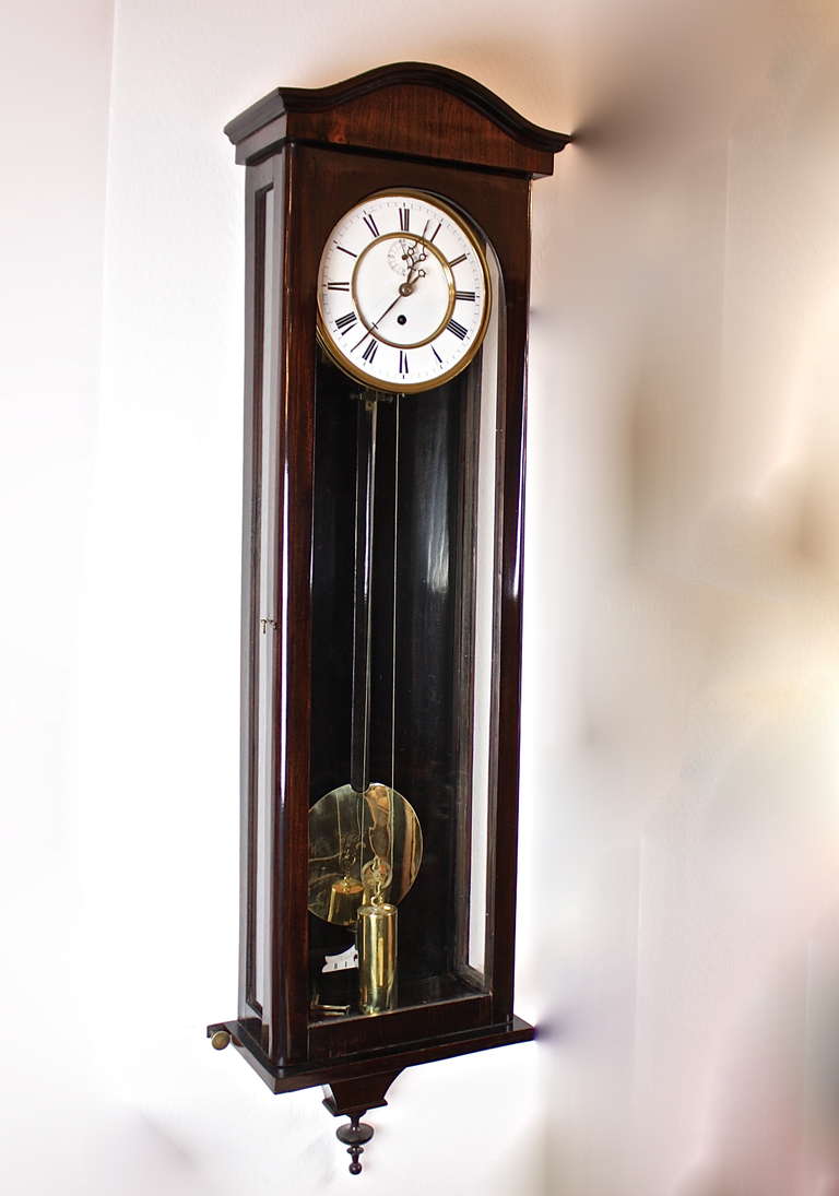 A Viennese ‘Dachluhr,’ the enamel dial in Roman numerals with subsidiary dials for seconds housed in an mahogany case with three glass panes. A brass pendulum and one brass weight. The dial held within a brass engine-turned bezel. The pediment