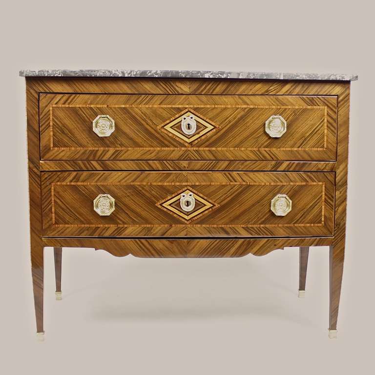 An Italian two-drawered Louis XVI commode, surmounted by a ‘Gris St Anne’ marble-top, on tall, square tapering legs in gilt bronze sabots, the front and sides veneered with crossbanding and a lozenge shaped veneered center, mounted with ribbon tied