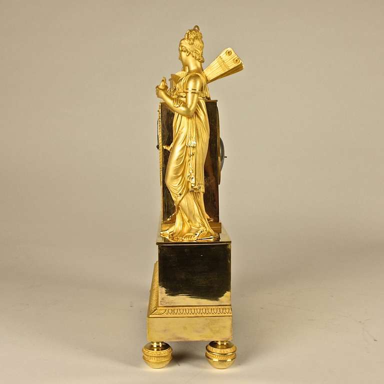 A fine French Empire mantle clock with a rectangular drum case surmounted by a classical urn, a treasure chest and a butterfly, flanked by a standing and winged figure of Psyche holding a Roman oil lamp and a dagger and by a column with a flamed