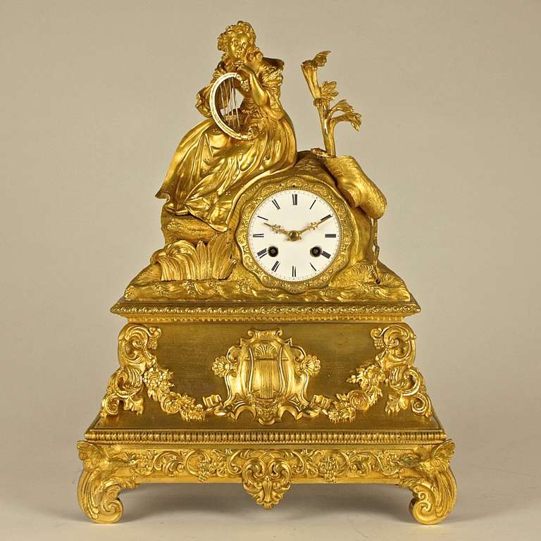 A fine gilt-bronze mantle clock, featuring a young woman playing the harp. Rich gilt decoration playing with matt and polished surfaces and intricate details extends to the weave of the hat and other details. 
The white enamel with Roman numerals
