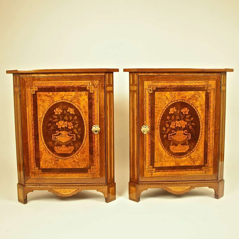An exceptional pair of corner cabinets each with a marquetry top, above  canted corners simulating fluting, the panelled door with flowers in a vase  made of precious woods i.e. burl ash, walnut, fruit woods, with pyrogravure  and fruitwood