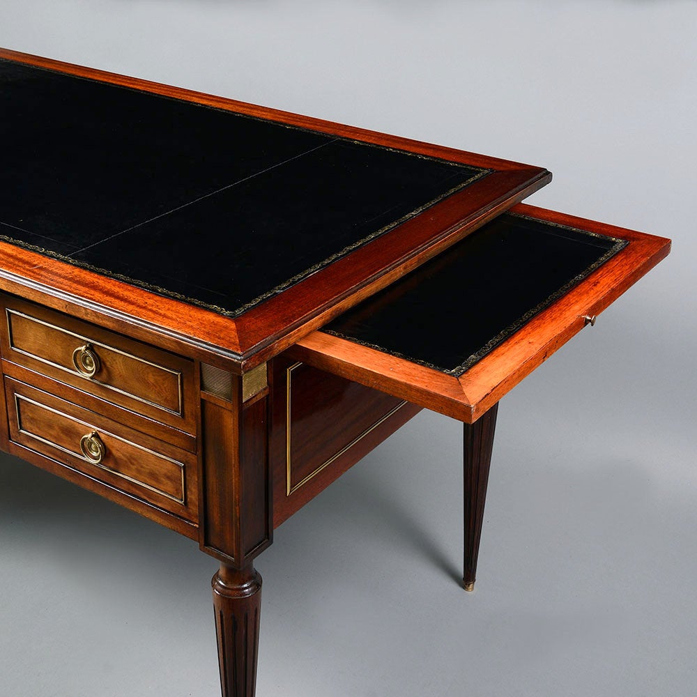 A late 19th century mahogany bureau plat or writing table, the cleated top with black leather inset, concealing side slides, all above one long drawer, flanked by two short drawers on either side, with brass medallion handles and rectangular panels,