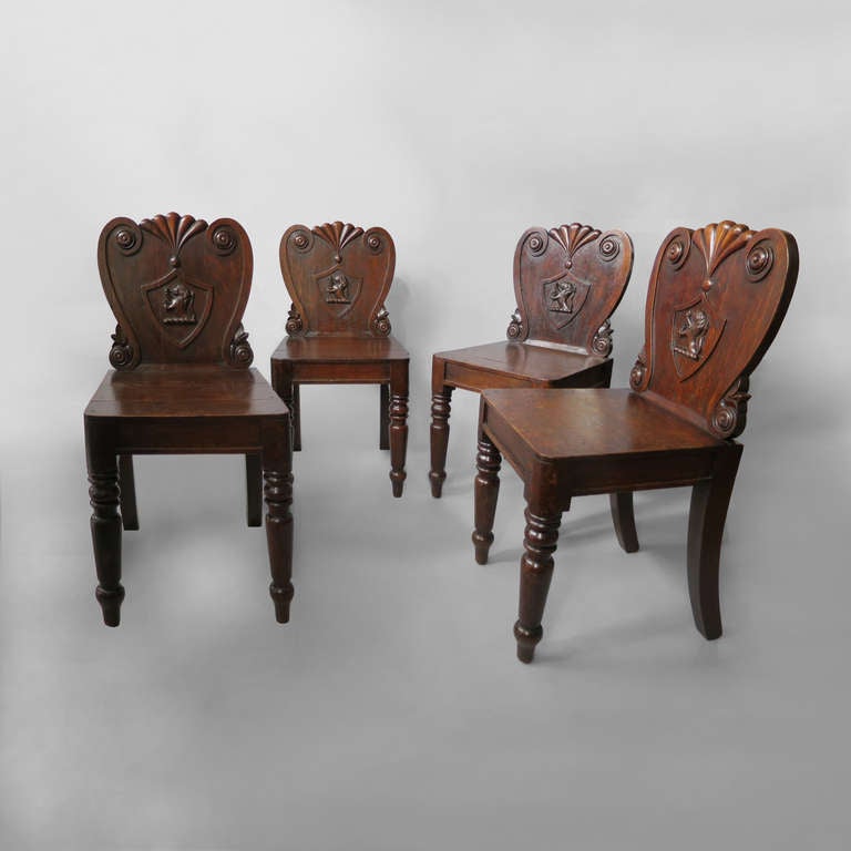 A set of four Regency Period oak hall chairs, the generously carved cartouche backs with gradooned crestings, scrolling edges and having turned front legs. The backs all depicting a crest: a lion rampant holding a trefoil.