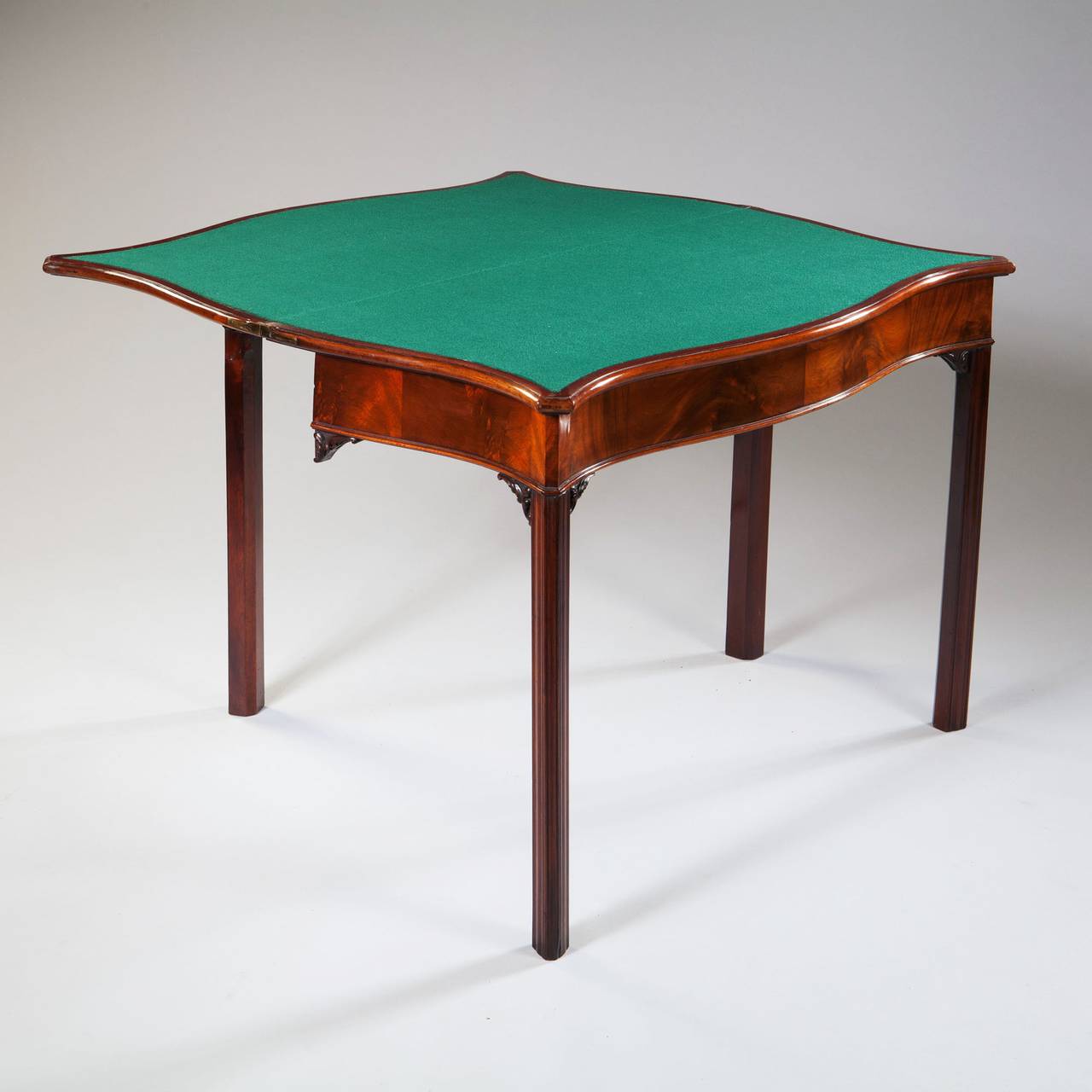 A George III Period mahogany card table, the finely figured flame veneered top of serpentine form, fitted with green baize playing surface, the chamfered square legs headed with carved rococo brackets.