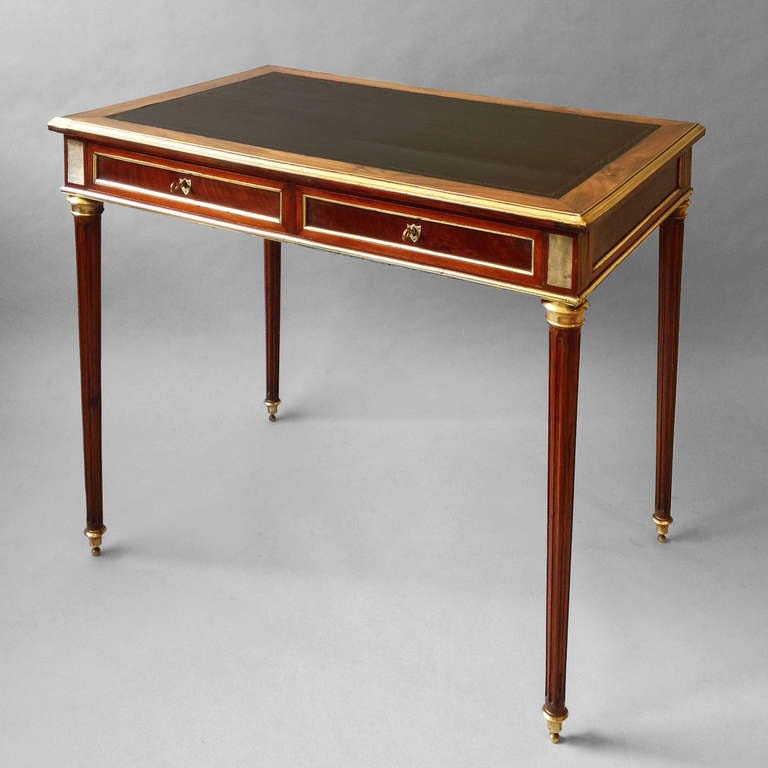 A late 19th century Louis XVI style writing table, the brass rimmed walnut top inset with black gilt tooled leather surface above two drawers with brass moulding and shield shaped escutcheons, the whole raised on turned fluted legs with brass feet.