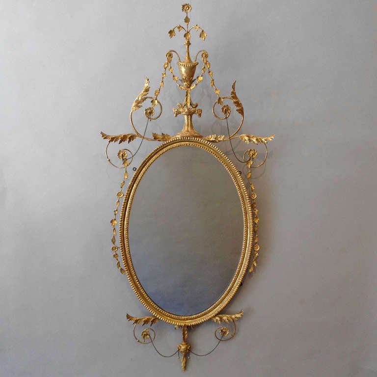 A late 18th Century carved giltwood mirror in the Adam taste, the gadrooned oval frame surrounded by carved floral details, a Classical urn, stylised acanthus and foliate carving and rams heads, all liked by chains with gilded drops.